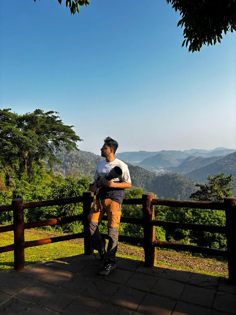 Ashwin Tak standing at a wooden railing overlooking a misty forested valley, holding a telephoto camera