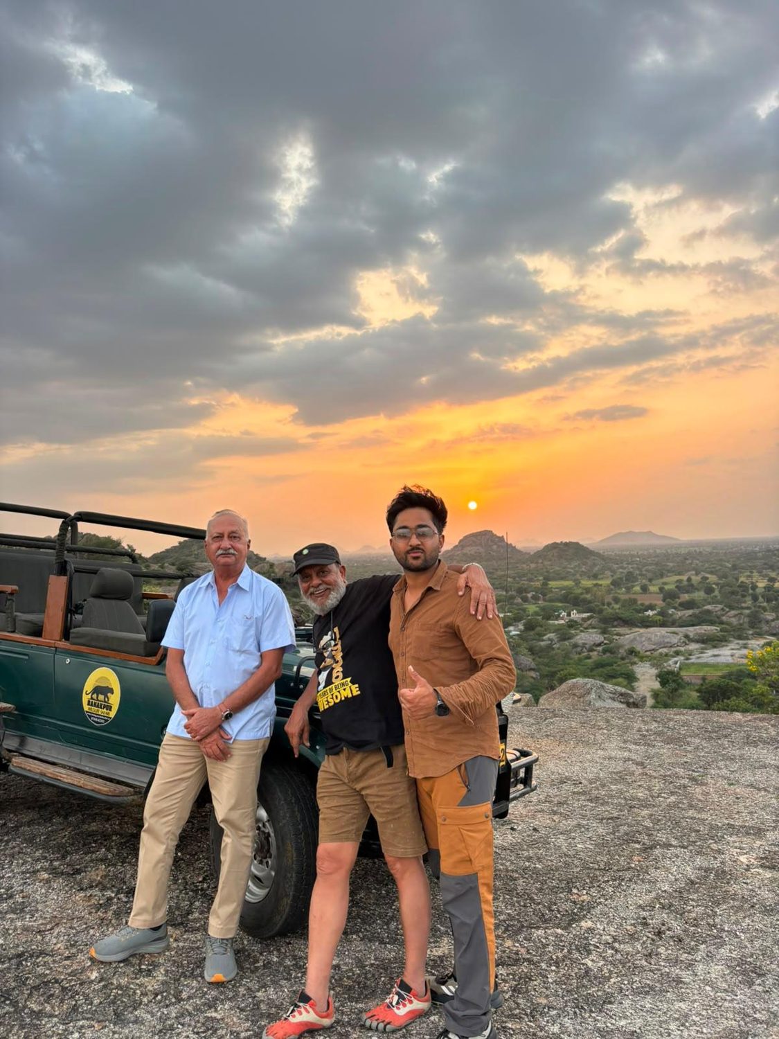 Three men standing beside a safari jeep on a rocky hilltop at sunset near Ranakpur